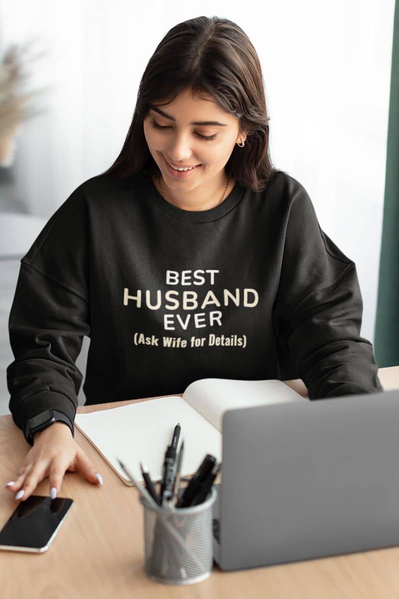 couples outfit sweatshirt with a woman wearing a black sweatshirt with humorous text, sitting at a desk with a laptop and phone.