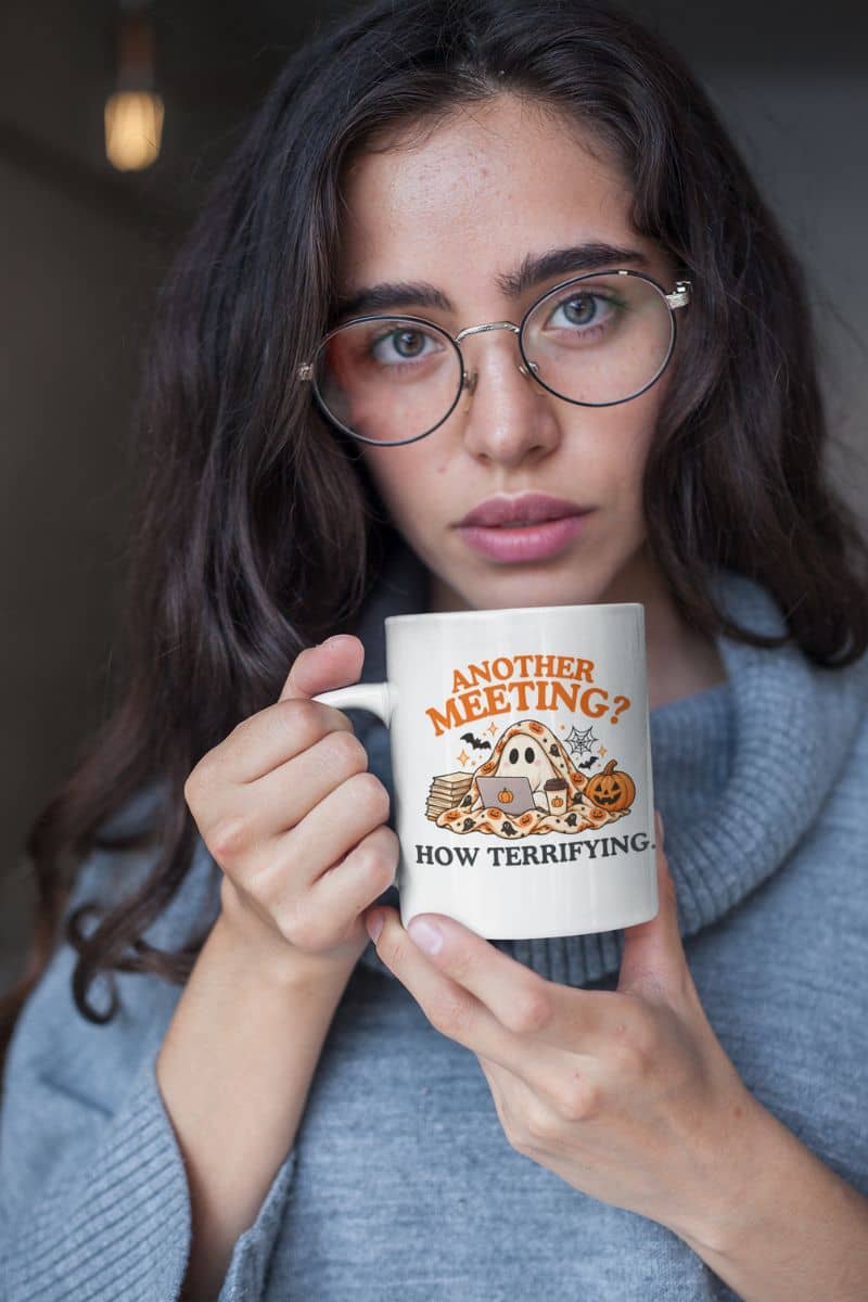 A woman with dark hair and round glasses holding a white ceramic mug featuring the “Another Meeting? How Terrifying.” design, inspired by humorous coworker coffee mugs.