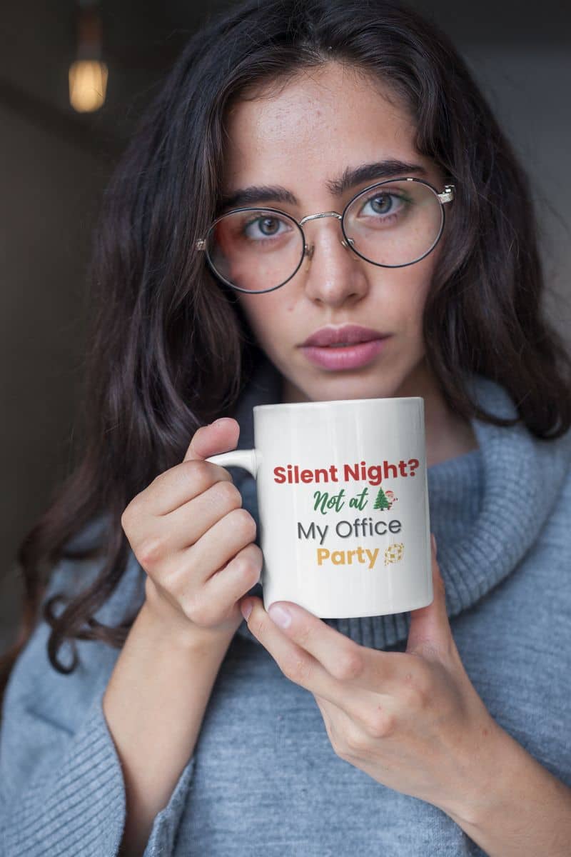 coworker holiday drinkware with a woman holding a mug with text 'Silent Night? Not at My Office Party', wearing glasses and a gray sweater.