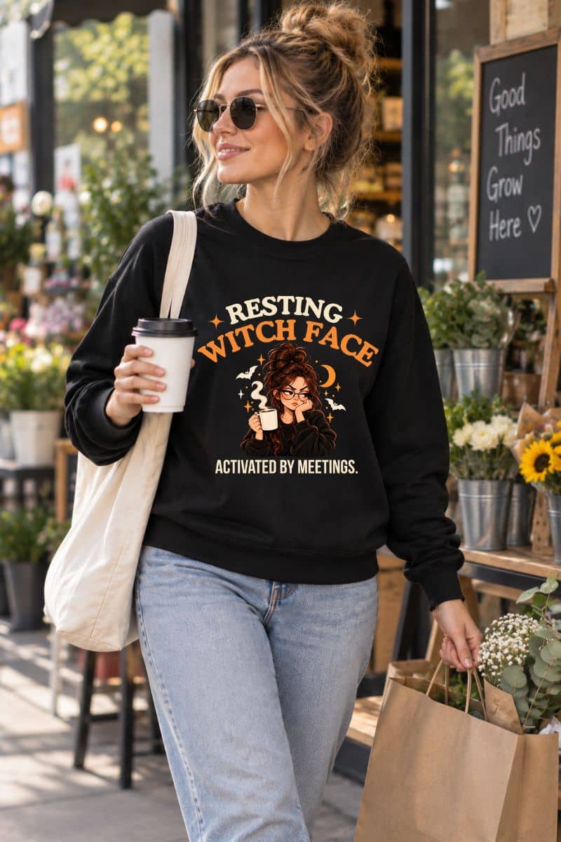 A woman walking outdoors wearing a sweatshirt featuring the text 'Resting Witch Face. Activated by Meetings.' with a woman's face.