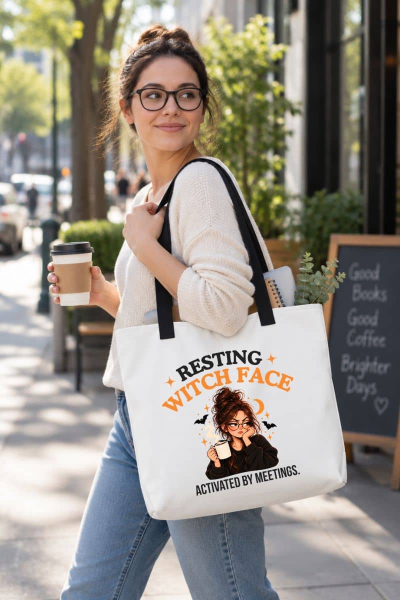 A woman walking outdoors on a city sidewalk carrying a large coworker tote bag with black handles and a printed graphic that reads 'Resting Witch Face. Activated by Meetings.' She is also holding a takeaway coffee cup.