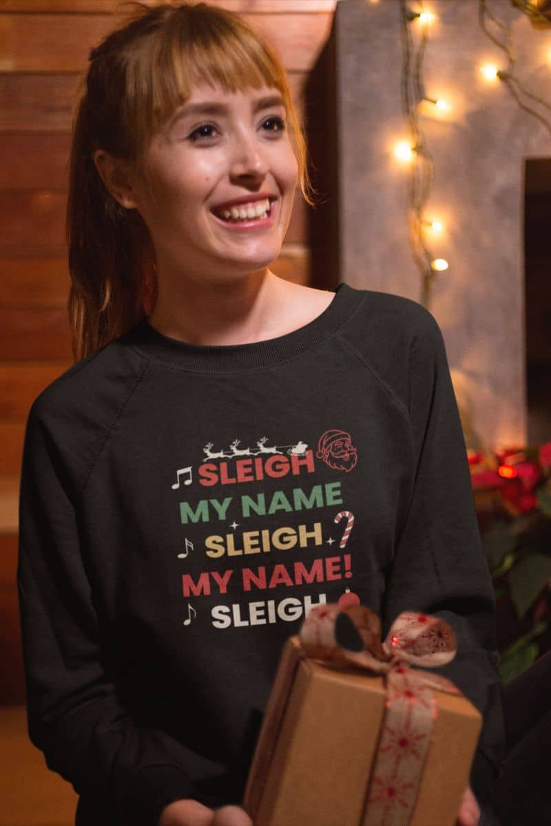 cozy Christmas outfit with a person wearing a black Christmas-themed sweater with text 'Sleigh My Name, Sleigh My Name', holding a gift box.