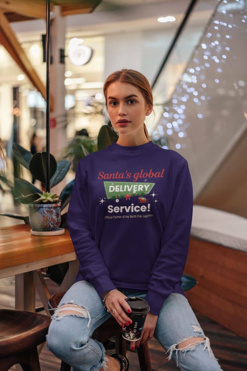 cozy Christmas sweater with a woman wearing a purple sweatshirt with text 'Santa's Global Delivery Service.' sitting in a modern indoor setting.
