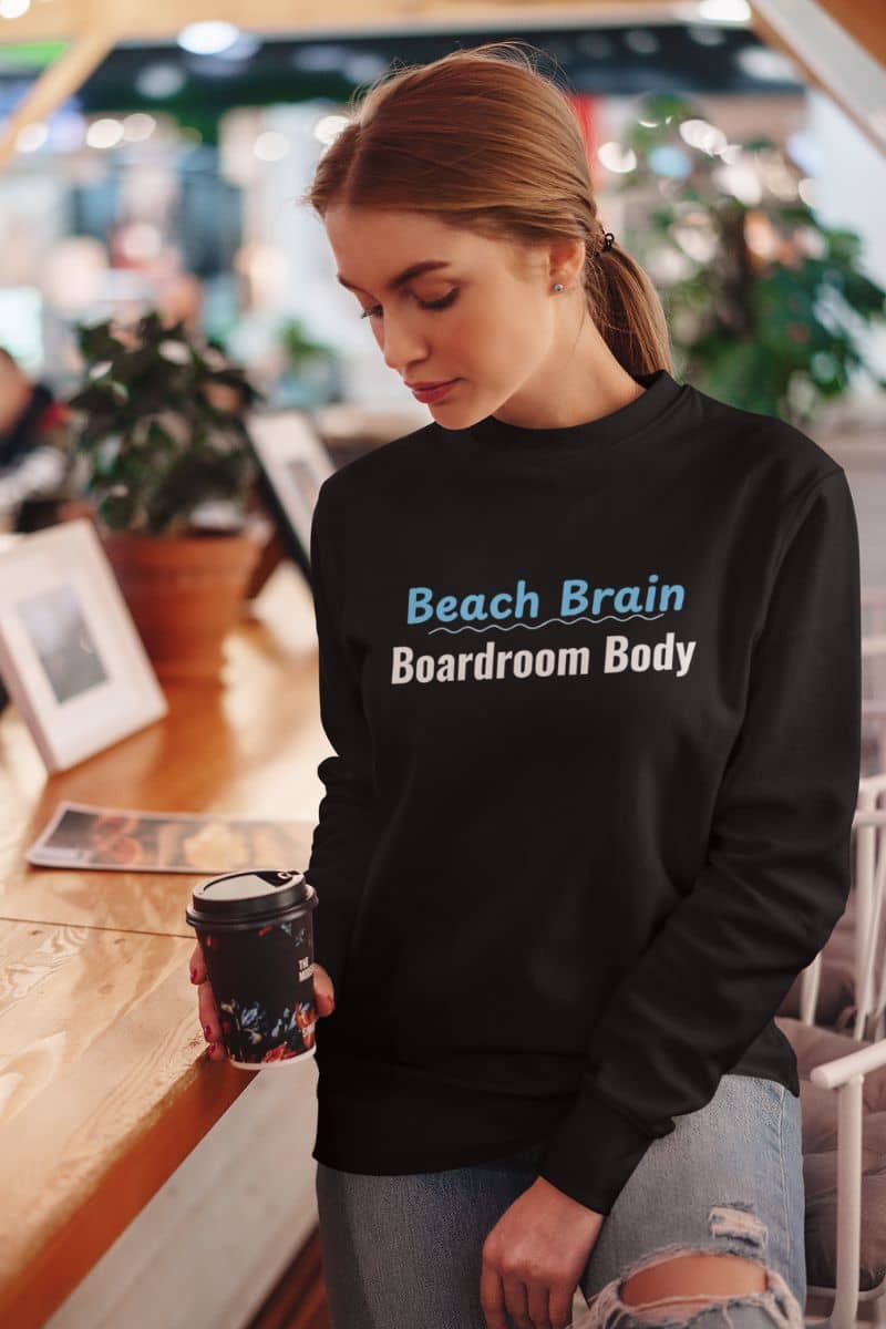 cozy beach sweatshirt with a woman wearing a black sweatshirt with 'Beach Brain Boardroom Body' text in a casual setting.