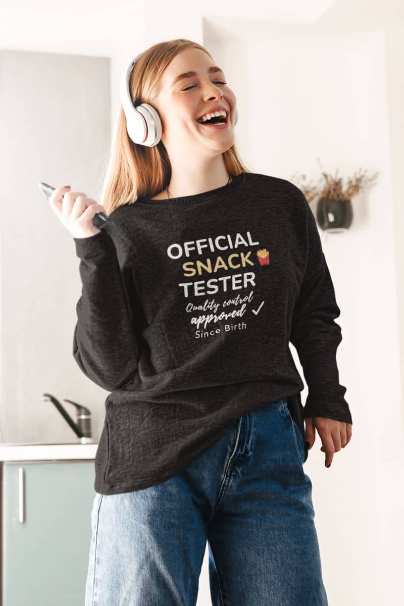 cozy casual sweatshirt with a person wearing a black sweatshirt with text 'Official Snack Tester – Quality Control Approved – Since Birth', standing in a kitchen.