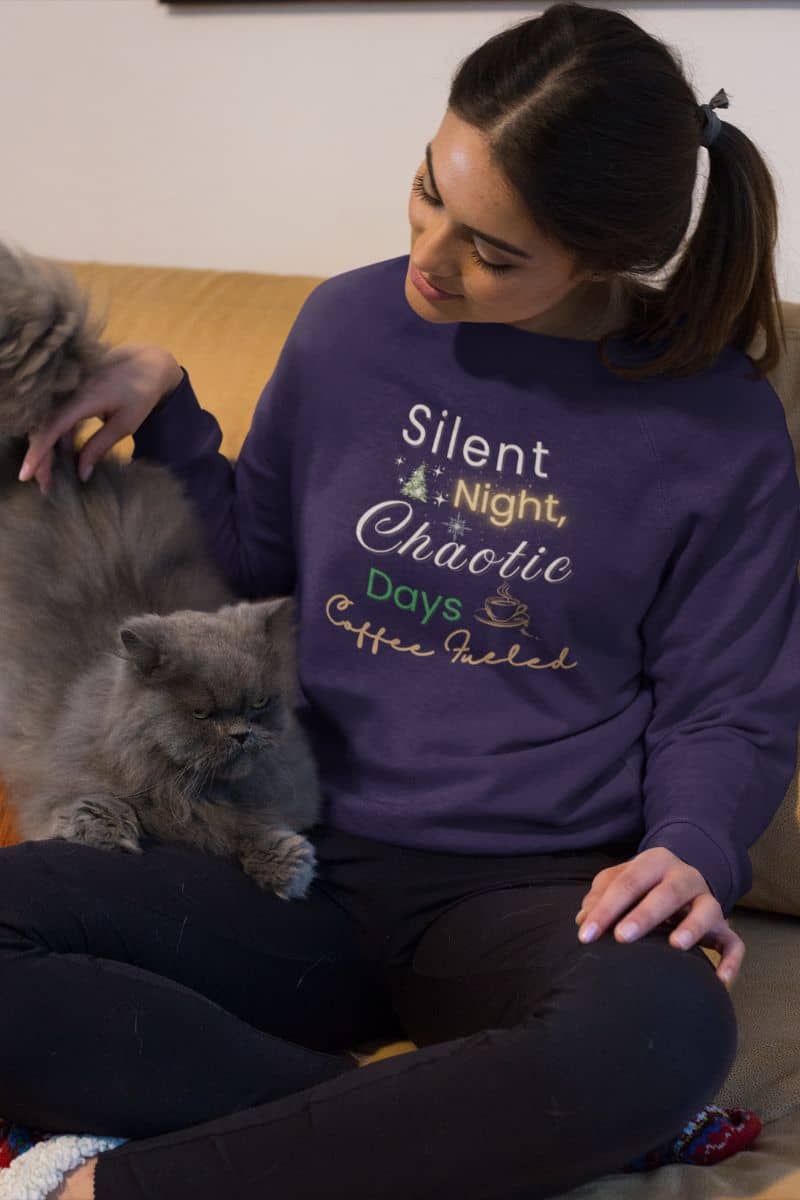 cozy christmas sweater with a woman wearing a purple sweatshirt with text 'Silent Night, Chaotic Days Coffee Fueled' sitting on a couch with two cats.