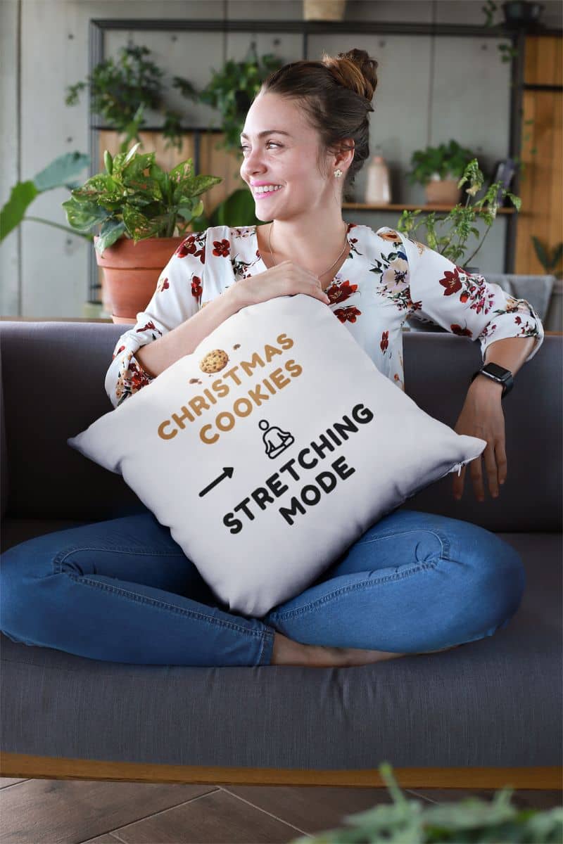 cozy christmas throw pillow with a woman sitting on a couch holding a pillow with text 'Christmas Cookies, Stretching Mode', surrounded by indoor plants.
