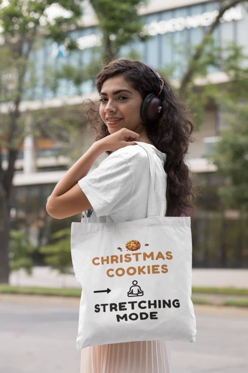 cozy christmas tote with a woman holding a tote bag with humorous text 'Christmas Cookies, Stretching Mode' outdoors