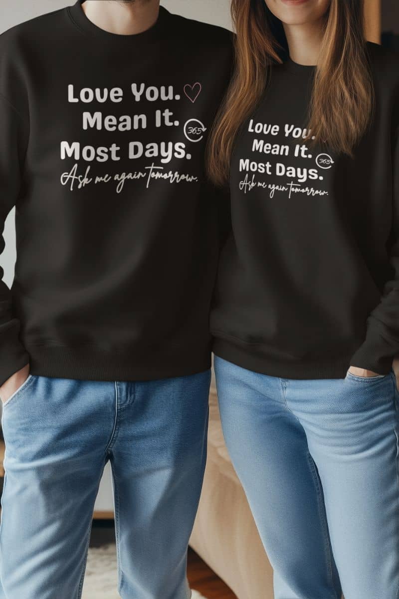 cozy couples humor top with two people wearing black sweatshirts with a humorous quote 'Love You. Mean It. Most Days. Ask me again tomorrow.', standing close together.