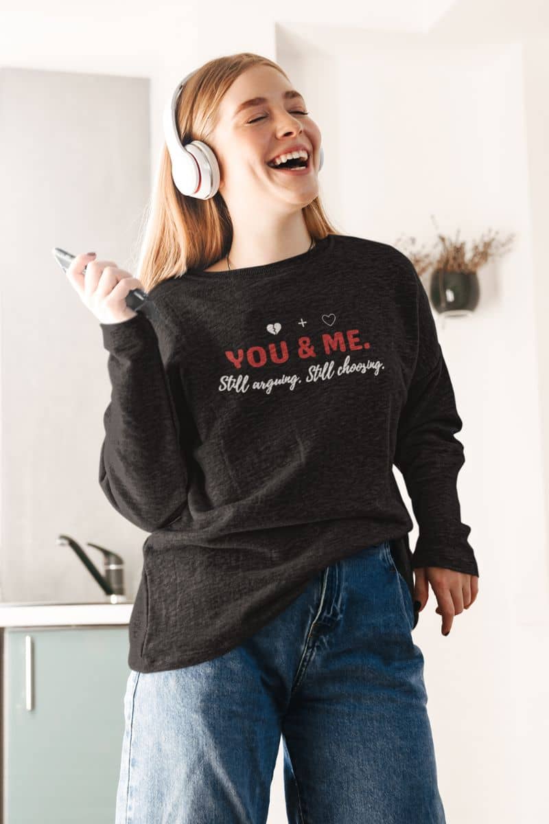 cozy couples sweatshirt with a person wearing a black sweatshirt with text 'YOU & ME. Still arguing. Still choosing.', laughing in a kitchen.