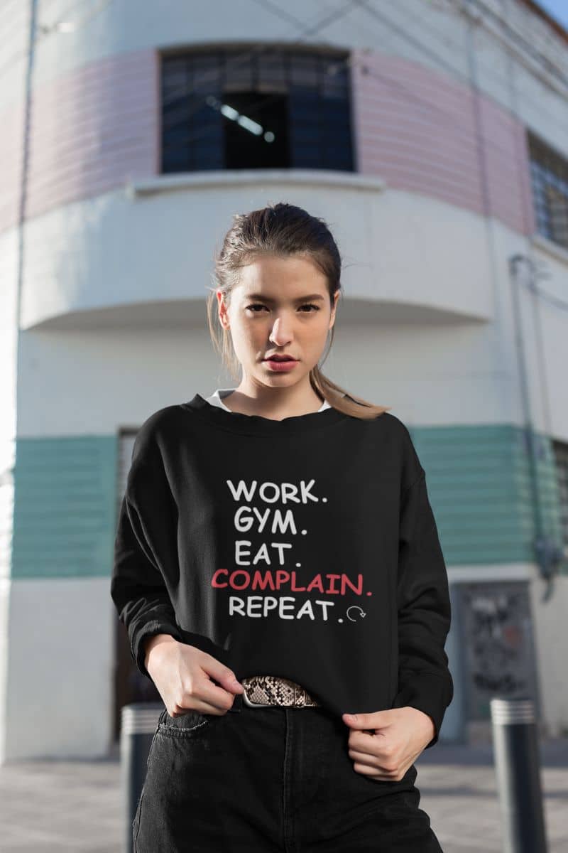 cozy everyday pullover with a woman in a black sweatshirt with text 'Work. Gym. Eat. Complain. Repeat' in an urban setting