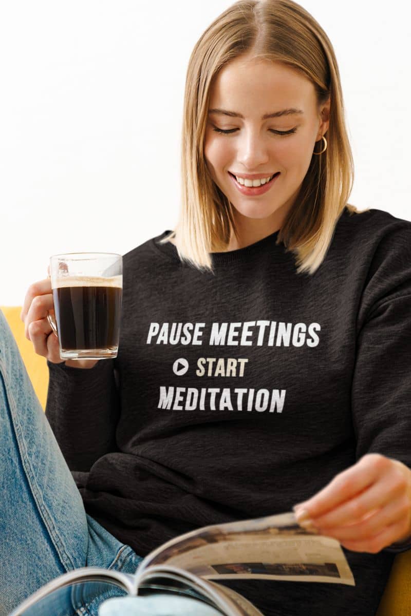 cozy meditation sweatshirt featuring a woman wearing a sweatshirt with the text 'Pause Meetings, Start Meditation', while reading a magazine and holding a cup of coffee