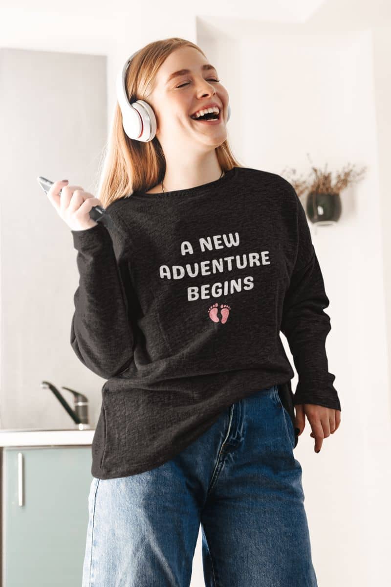 cozy parent pullover with a person wearing a black sweatshirt with 'A New Adventure Begins' text, holding a phone, in a kitchen setting.
