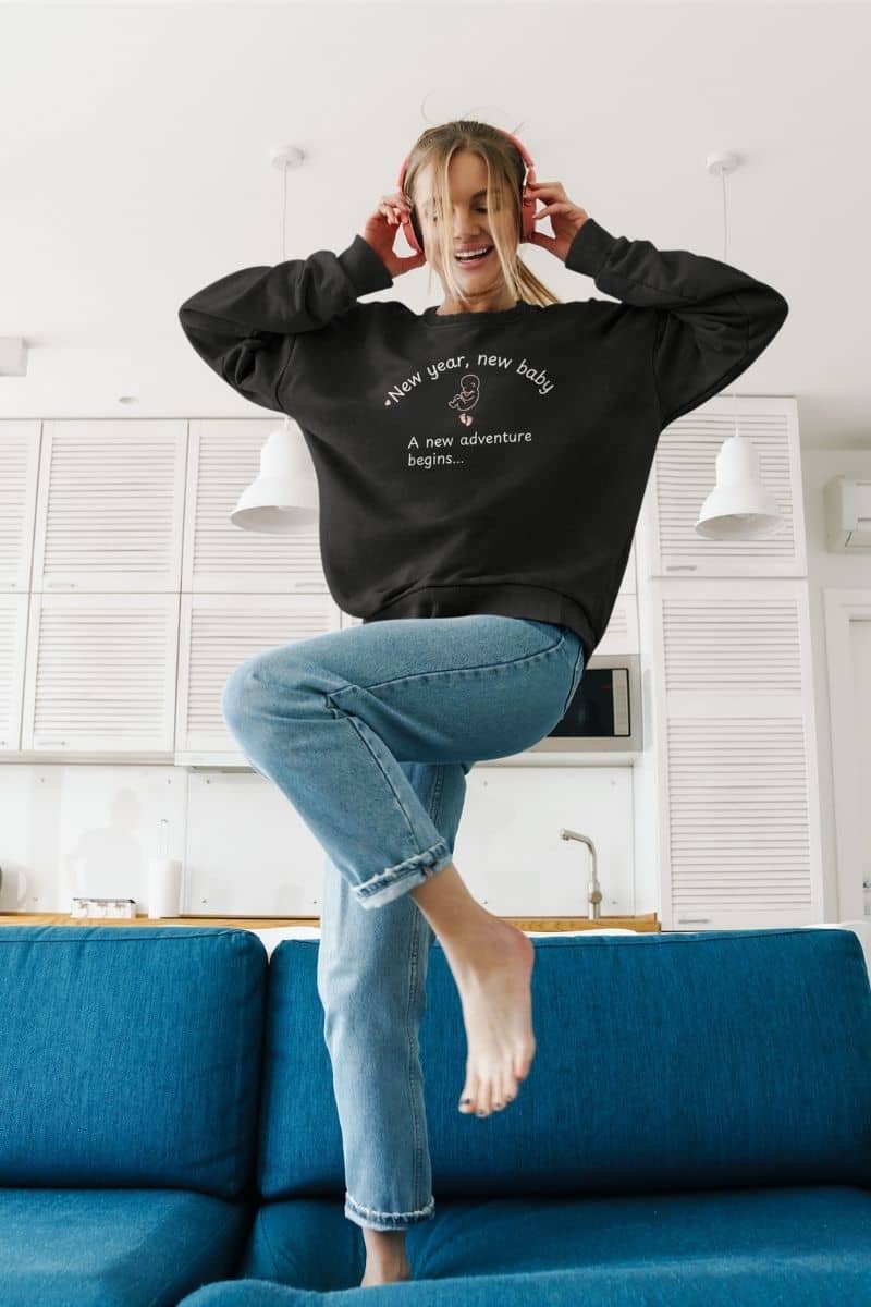 A cozy parent sweatshirt worn by a woman, featuring the text “New year, new baby. A new adventure begins…”, dancing on the couch while listening to music in the living room.