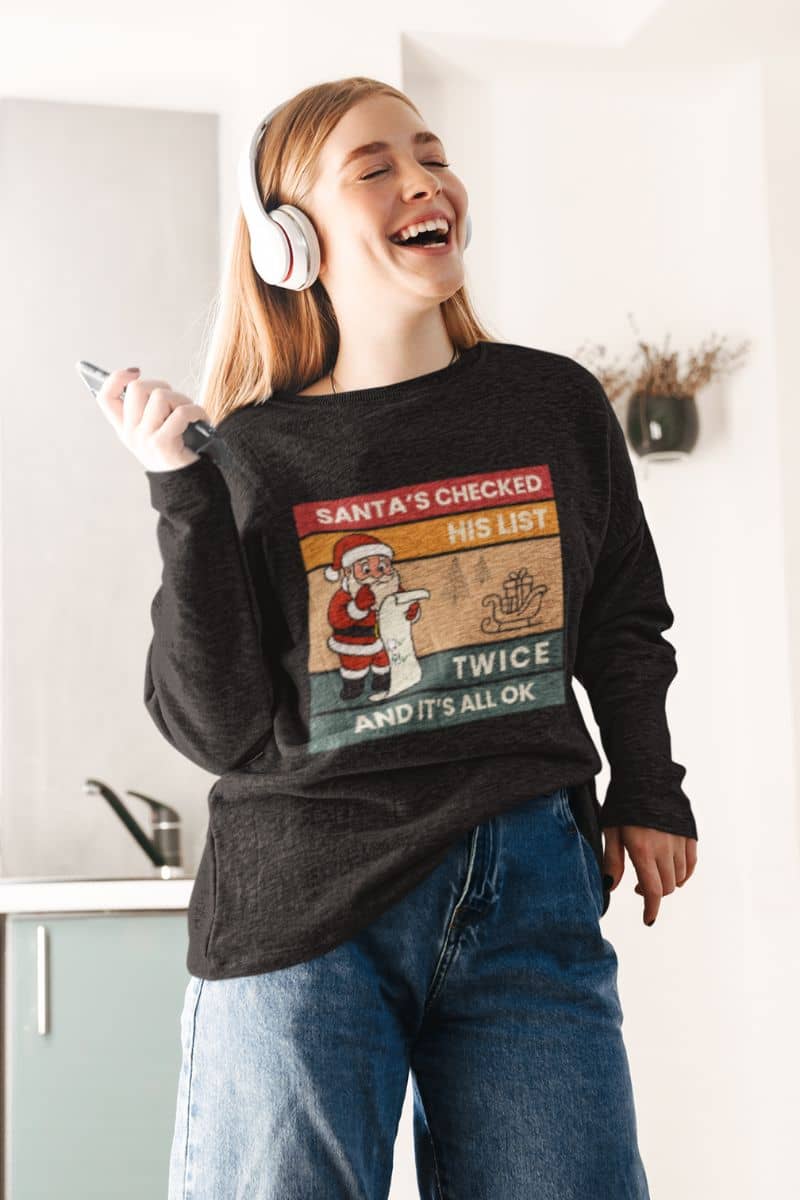 cozy santa sweatshirt with a person wearing a black sweater with text 'Santa's Checked His List Twice And It's All Ok', standing in a kitchen.
