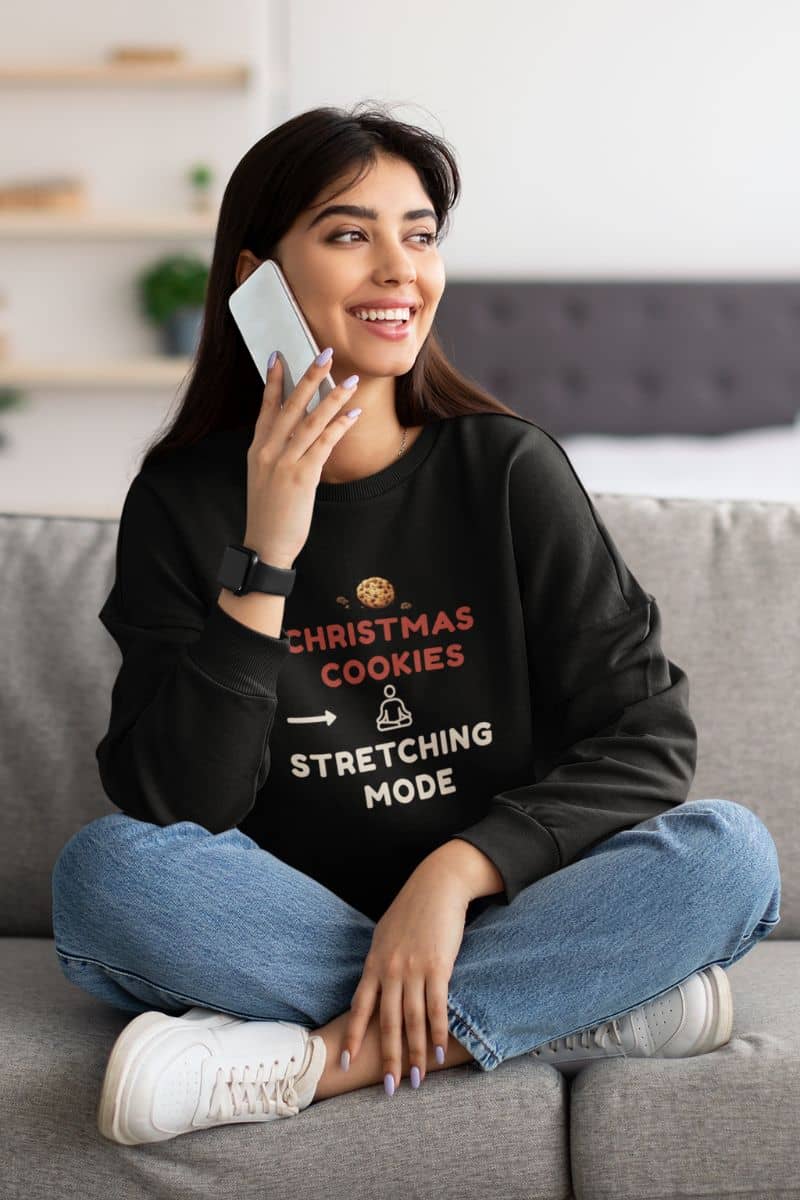 cozy self care sweatshirt with a woman sitting on a couch wearing a black sweatshirt with text 'Christmas Cookies, Stretching Mode', holding a phone.