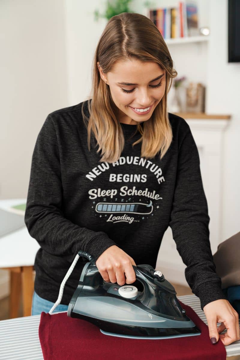 cozy tired life sweater with a woman ironing a shirt wearing a black sweatshirt with text 'New Adventure Begins, Sleep Schedule Loading…'.