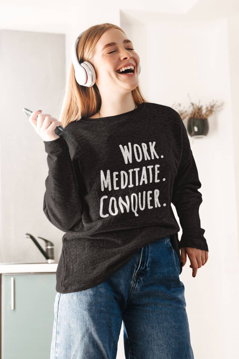 cozy work from home sweatshirt with a person wearing a black sweatshirt with 'Work. Meditate. Conquer.' text, laughing in a kitchen.