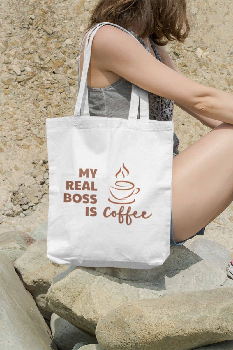 custom printed tote bags with a person holding a tote bag with 'My Real Boss is Coffee' text against a stone wall.