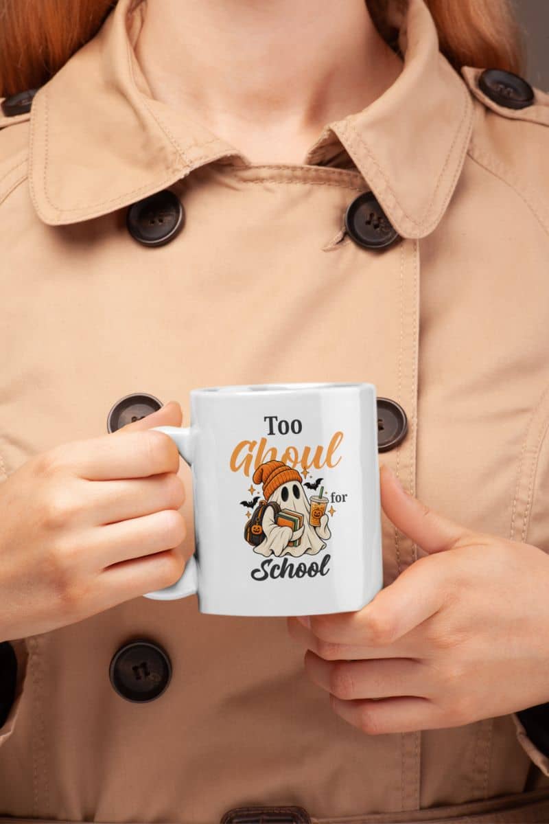 A person wearing a beige trench coat holding a cute Halloween mug featuring the text 'Too Ghoul for School' in orange and black lettering.
