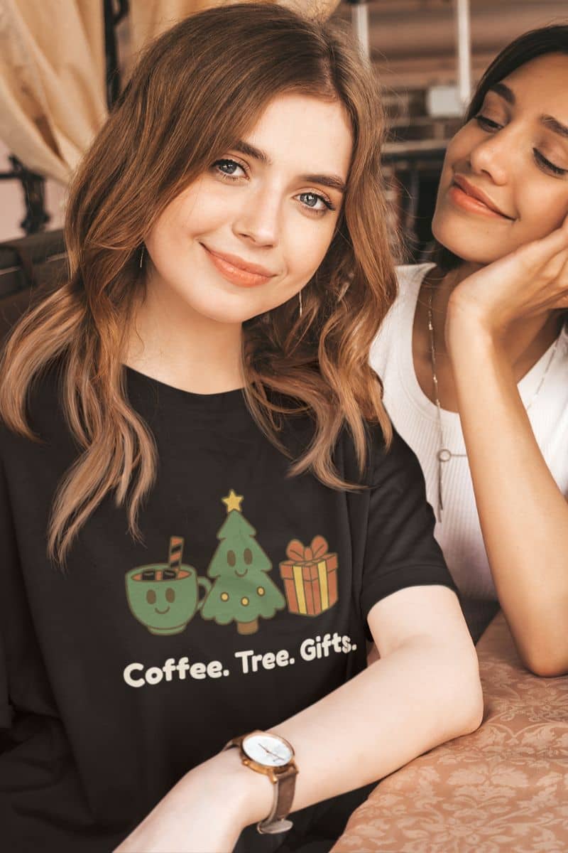 cute christmas graphic tee with a woman wearing a black t-shirt with Christmas-themed graphics and text 'Coffee. Tree. Gifts.', sitting next to another person.