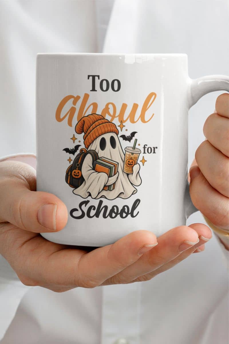 A person holding a cute ghost mug with a Halloween-themed graphic that reads 'Too Ghoul for School' in orange and black text, featuring a ghost holding books and coffee