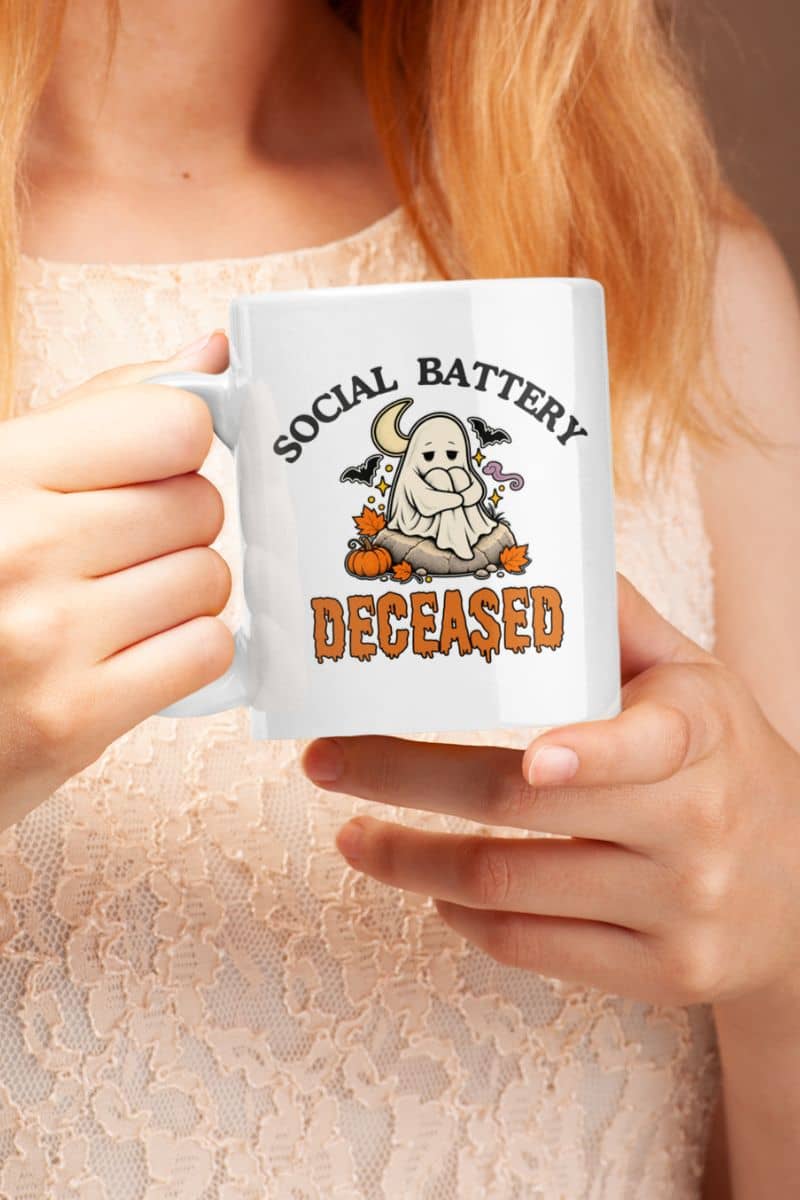 A person holding a cute ghost mug with a Halloween-themed graphic that reads 'Social Battery: Deceased' with a ghost, bats, and pumpkins.