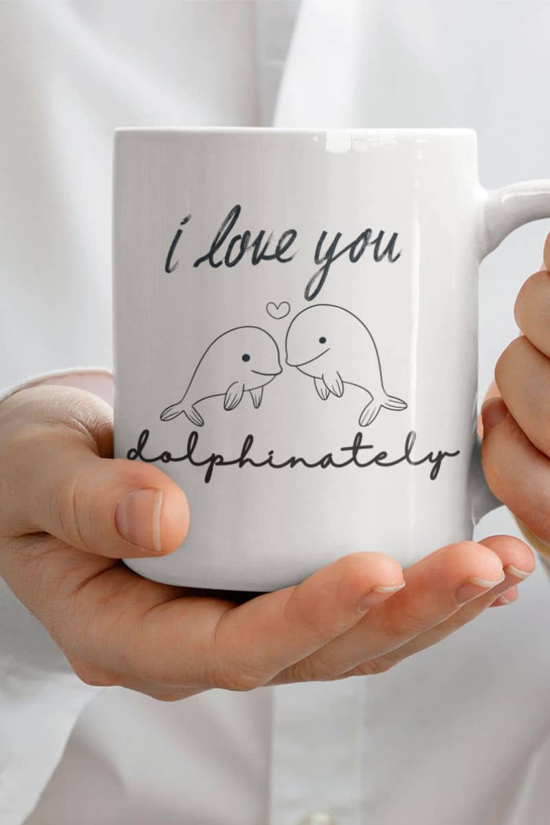 cute love coffee mug with a white mug with 'I love you dolphinately' text and dolphin illustrations held by hands.