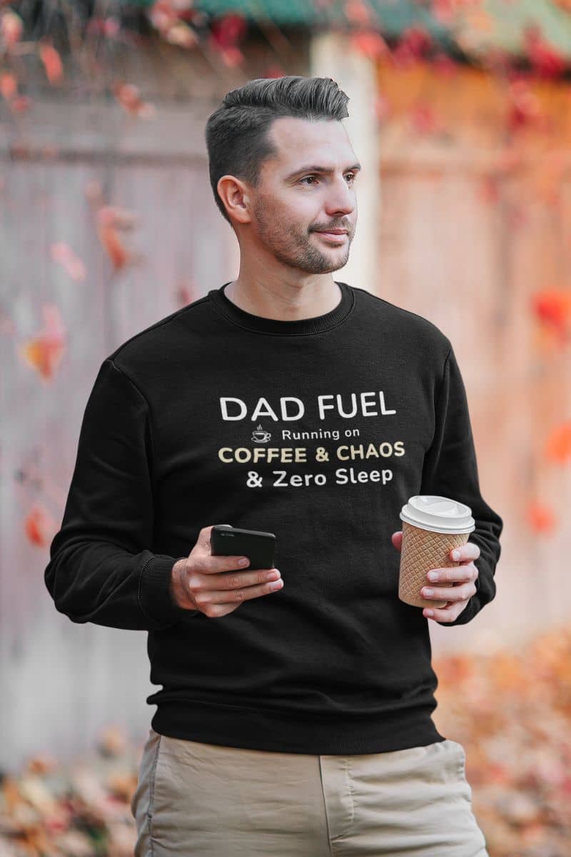 dad bod sweatshirt with a man wearing a black sweatshirt featuring the text 'DAD FUEL – Running on Coffee & Chaos & Zero Sleep', holding a coffee cup and phone, with a blurred autumn background.