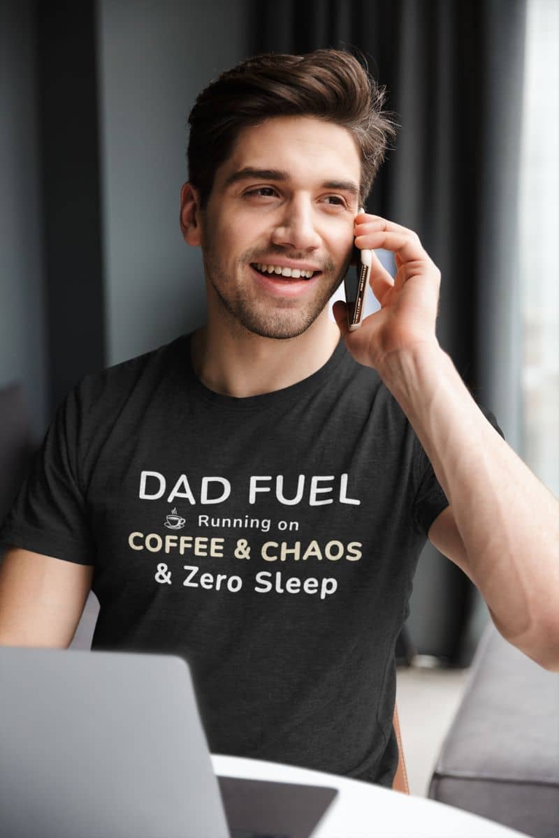 dad funny shirts with a man wearing a black t-shirt with 'DAD FUEL – Running on Coffee & Chaos & Zero Sleep' text, talking on a phone indoors.