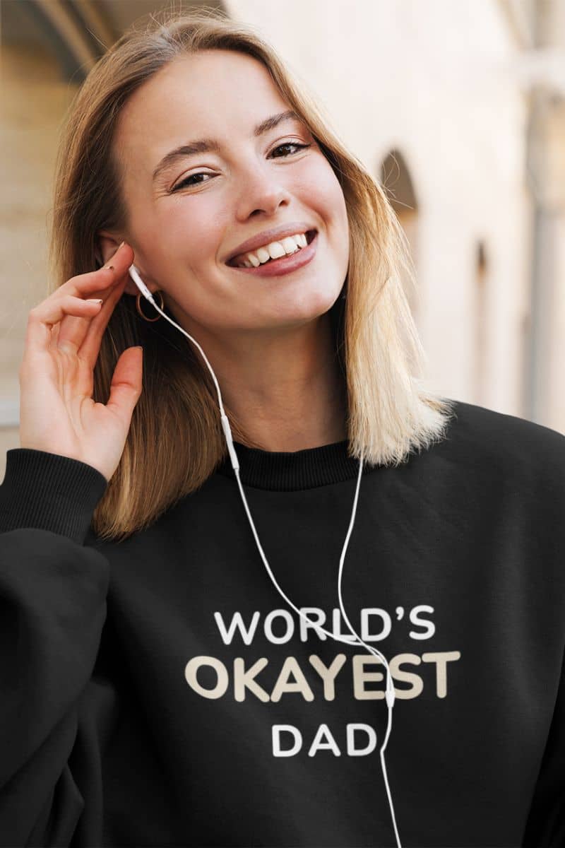 dad humor pullover with a person wearing a black sweatshirt with 'World's Okayest Dad' text, listening to music with earphones.