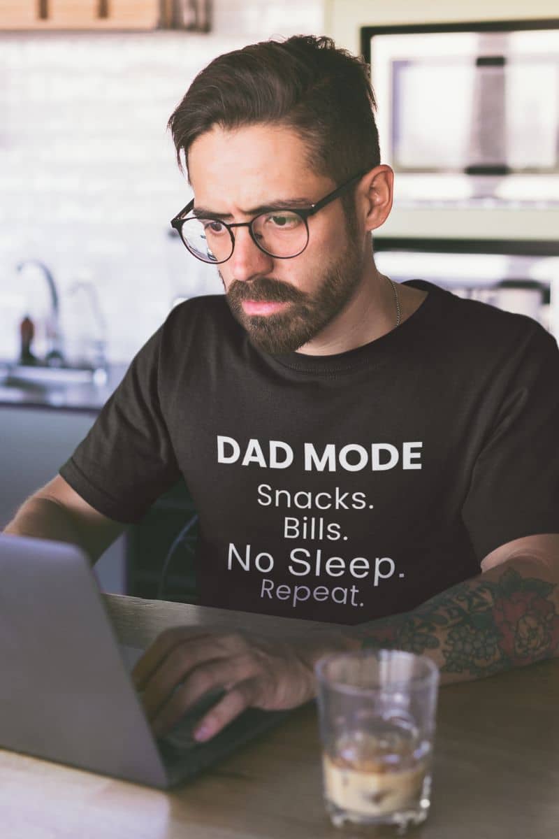 dad humor shirt with a man wearing a black t-shirt with text 'DAD MODE:  Snacks. Bills. No Sleep. Repeat.', sitting at a desk with a laptop.