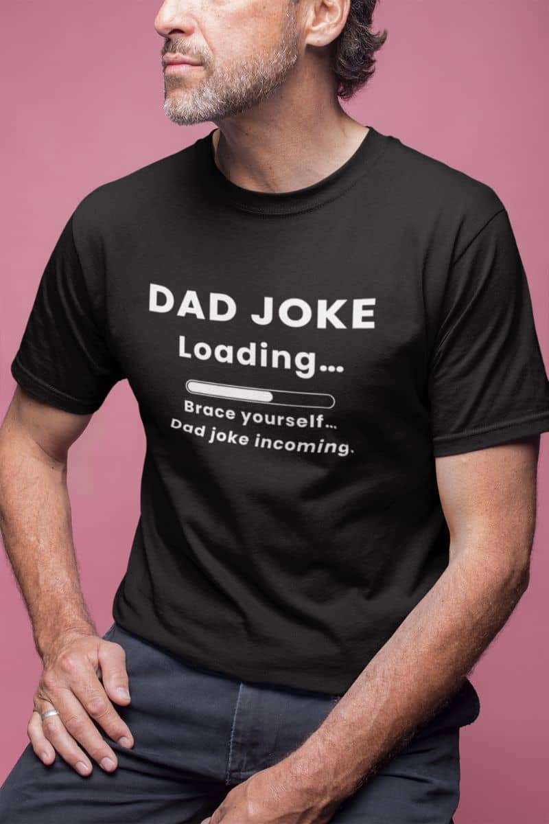 dad humor shirt with a man wearing a black t-shirt with text 'Dad Joke Loading, Brace yourself…Dad joke incoming.' on a pink background