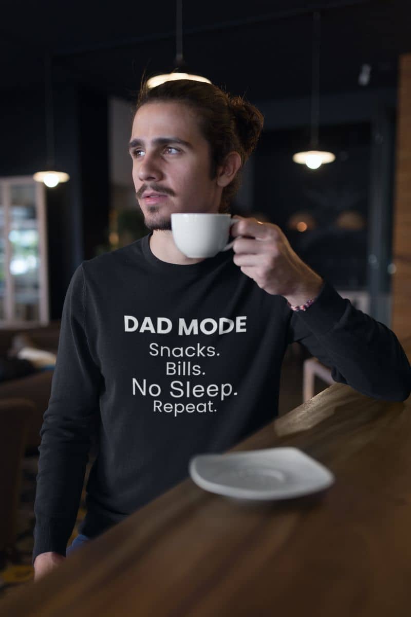 dad humor sweatshirt with a man holding a coffee cup in a bar wearing a black sweatshirt with 'DAD MODE:  Snacks.  Bills. No Sleep. Repeat.' text.