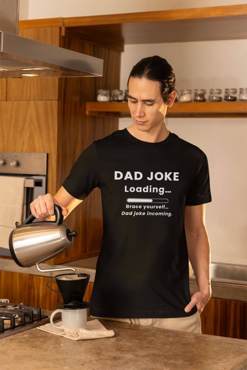 dad joke t shirt with a person in a kitchen wearing a black t-shirt with text 'Dad Joke Loading, Brace yourself…Dad joke incoming.', pouring coffee.