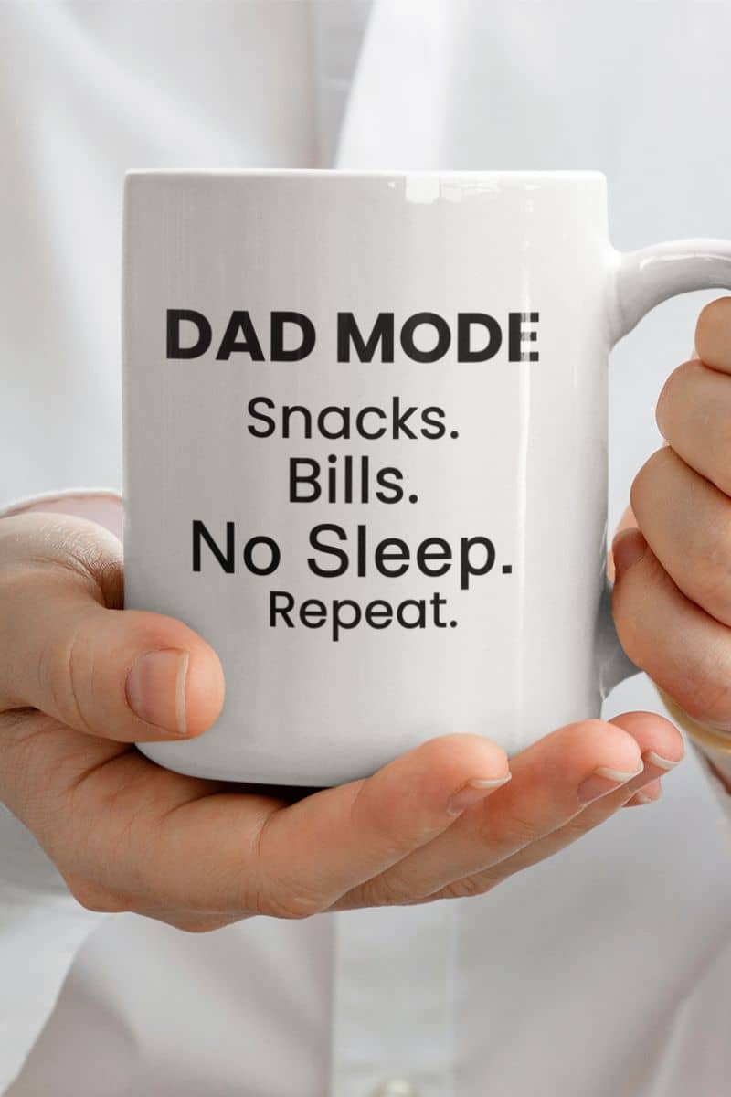 dad life coffee mug with a white mug with text 'DAD MODE: Snacks. Bills. No Sleep. Repeat.' held by a person.
