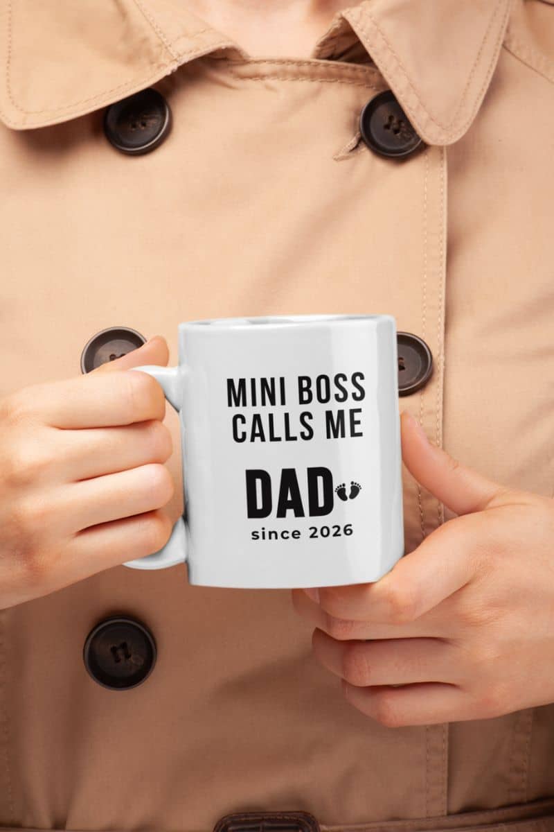 dad to be mug with a person holding a mug with text 'Mini Boss Calls Me Dad since 2026', wearing a beige coat.