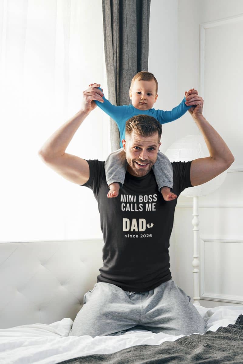 dad to be shirt with a man wearing a black t-shirt with text 'Mini boss calls me Dad since 2026', holding a child on his shoulders in a bedroom.