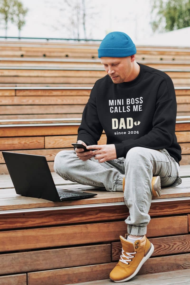 dad to be sweatshirt with a man sitting on wooden steps using a laptop and phone, wearing a black sweatshirt with text 'Mini Boss Calls Me Dad – since 2026'.
