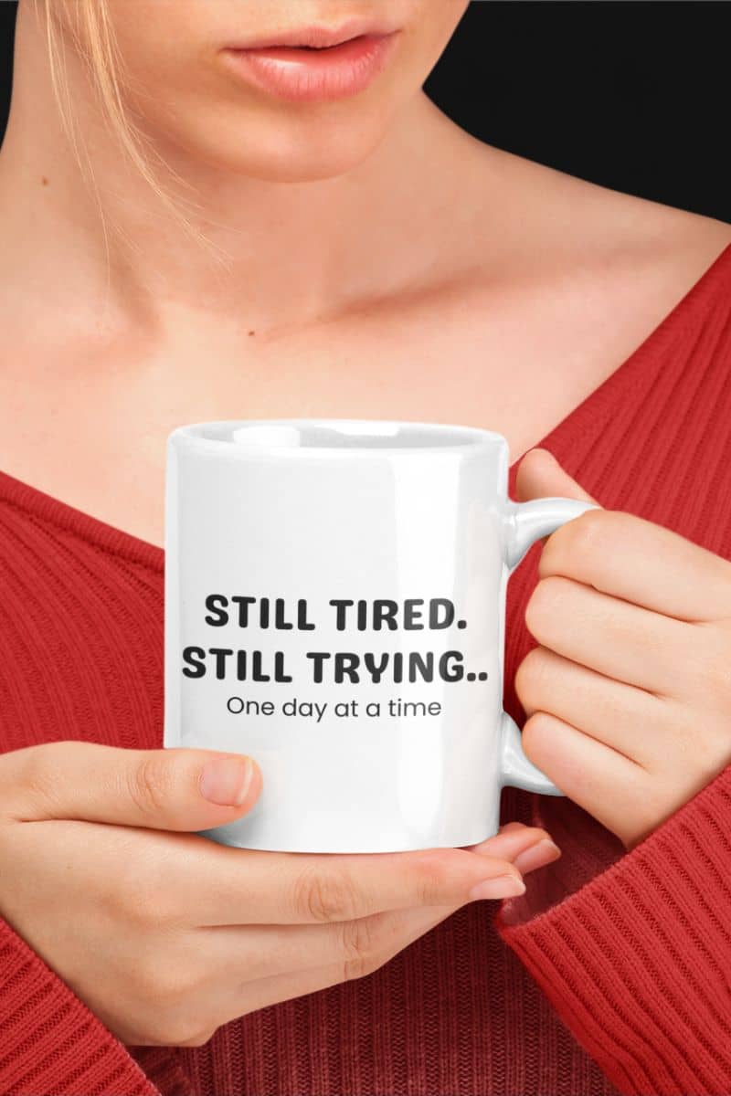 daily affirmation mug with a person holding a white mug with text 'Still Tired. Still Trying.. One day at a time' against a black background.