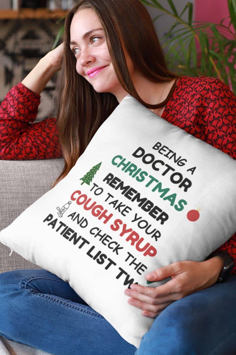 doctor holiday pillow with a woman holding a pillow features the phrase 'Being a doctor during Christmas, remember to take your cough syrup and check the patient list twice', sitting on a couch.