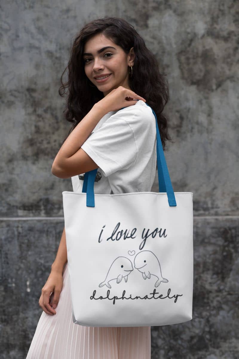 dolphin love tote bag with a woman holding a tote bag with 'I love you dolphinately' text and dolphin illustration against a stone wall.