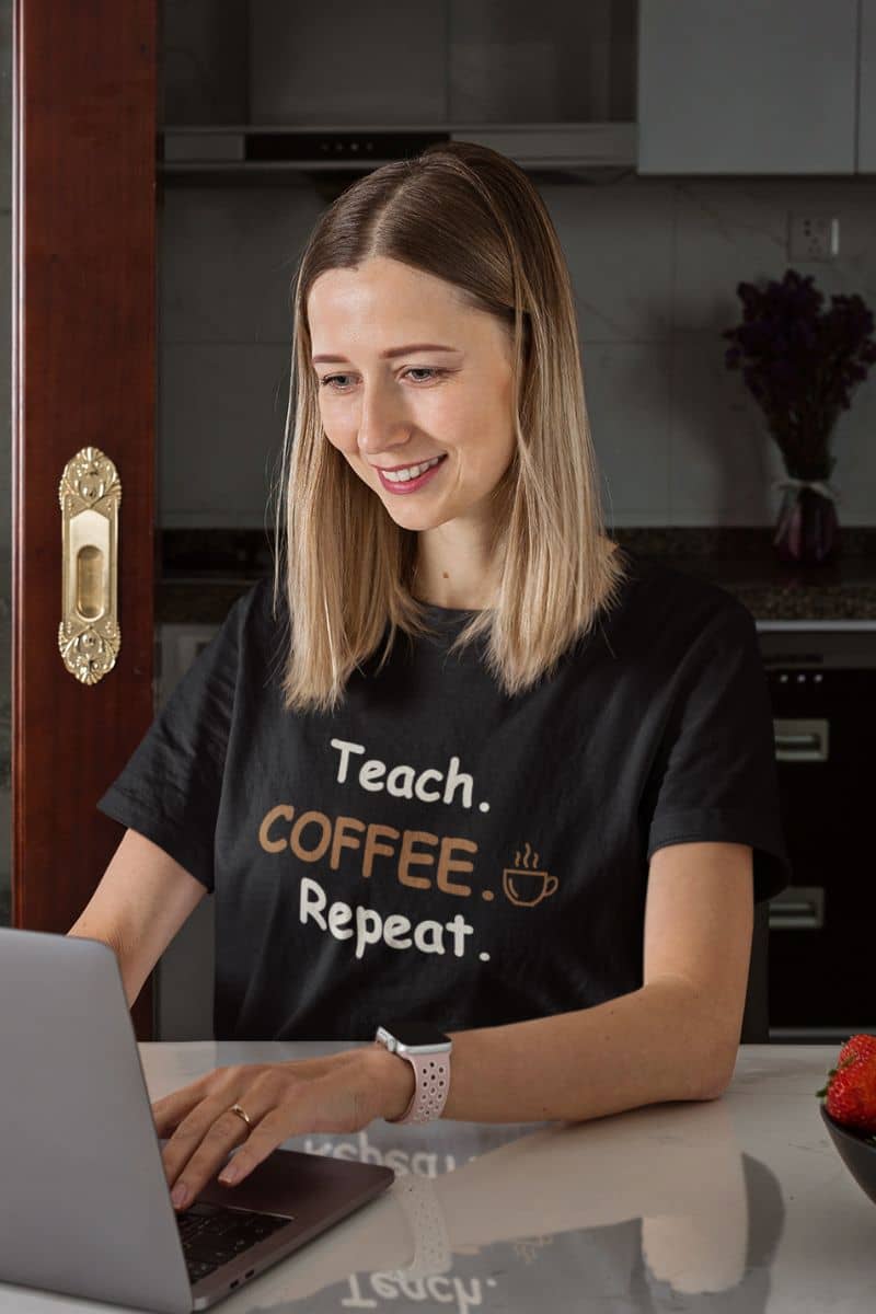 educator shirt with a woman with blonde-highlighted hair sits at a kitchen counter using a laptop, wearing a black graphic t-shirt that reads 'Teach. COFFEE Repeat.