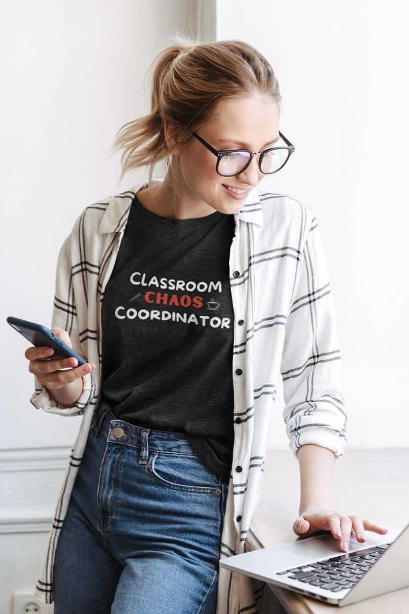elementary teacher shirt with a woman wearing glasses, a white plaid button-up shirt, and a black graphic t-shirt that reads 'Classroom Chaos Coordinator', holding a smartphone and using a laptop.