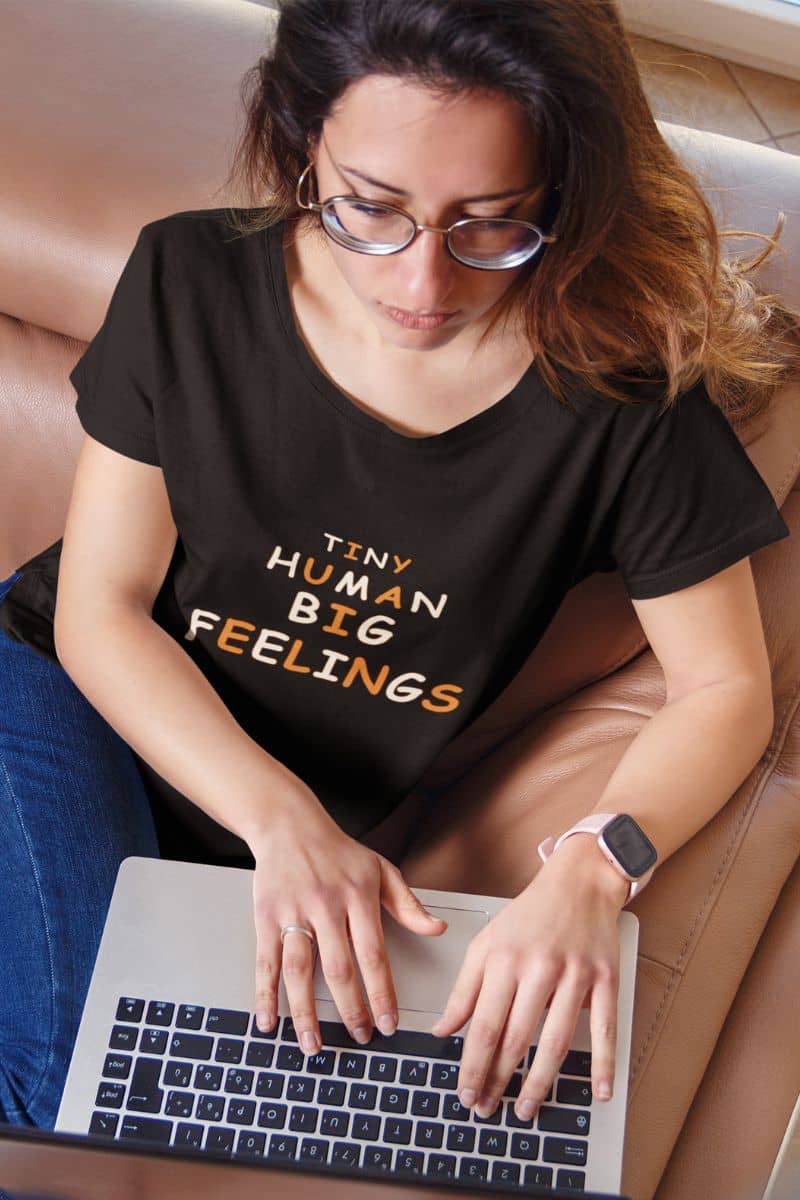 elementary teacher shirt with a woman wearing a black graphic t-shirt with the text 'Tiny Human Big Feelings' in white and orange, sitting on a tan leather couch and typing on a laptop.
