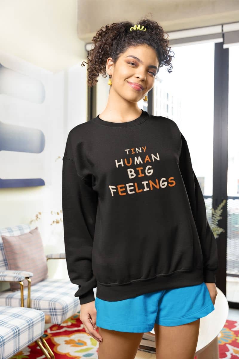 elementary teacher sweater with a woman wearing a black sweatshirt featuring the text 'TINY HUMAN BIG FEELINGS' in white and orange lettering, paired with bright blue shorts, standing in a modern living room setting.