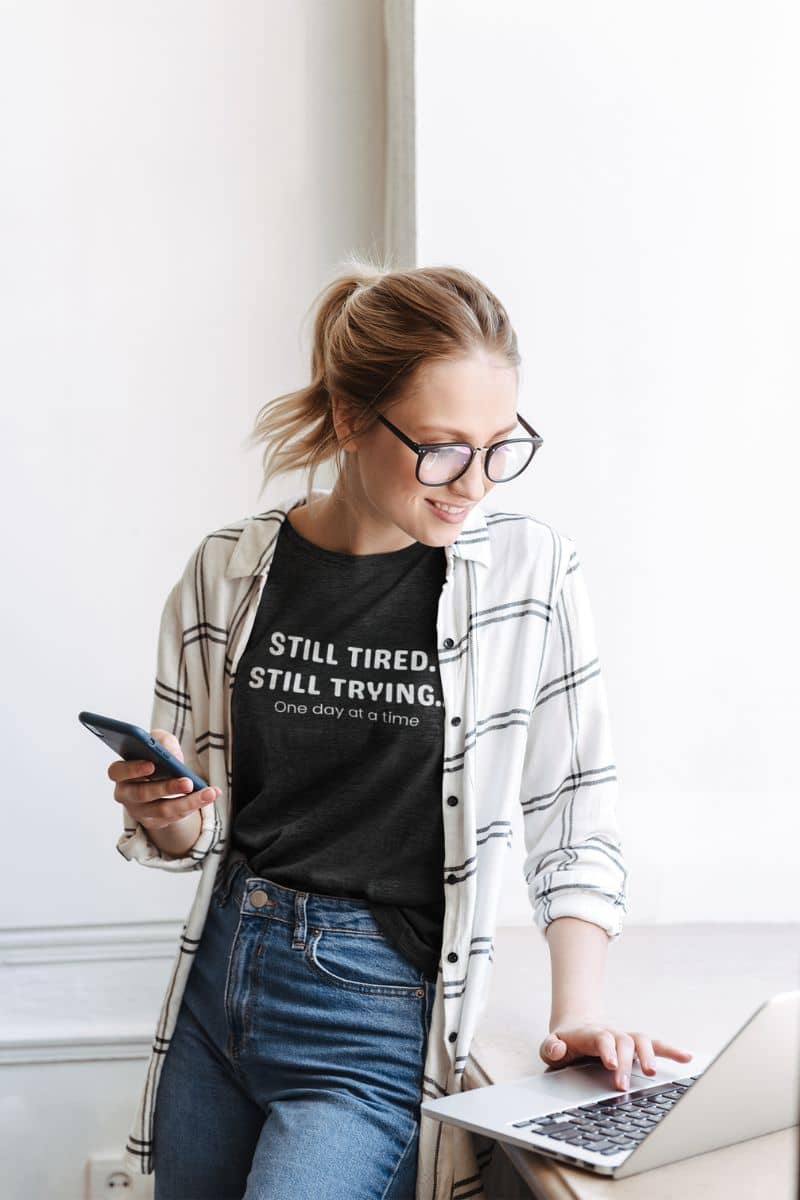emotional wellness shirt with a woman wearing a black t-shirt with 'STILL TIRED. STILL TRYING' text, using a laptop.