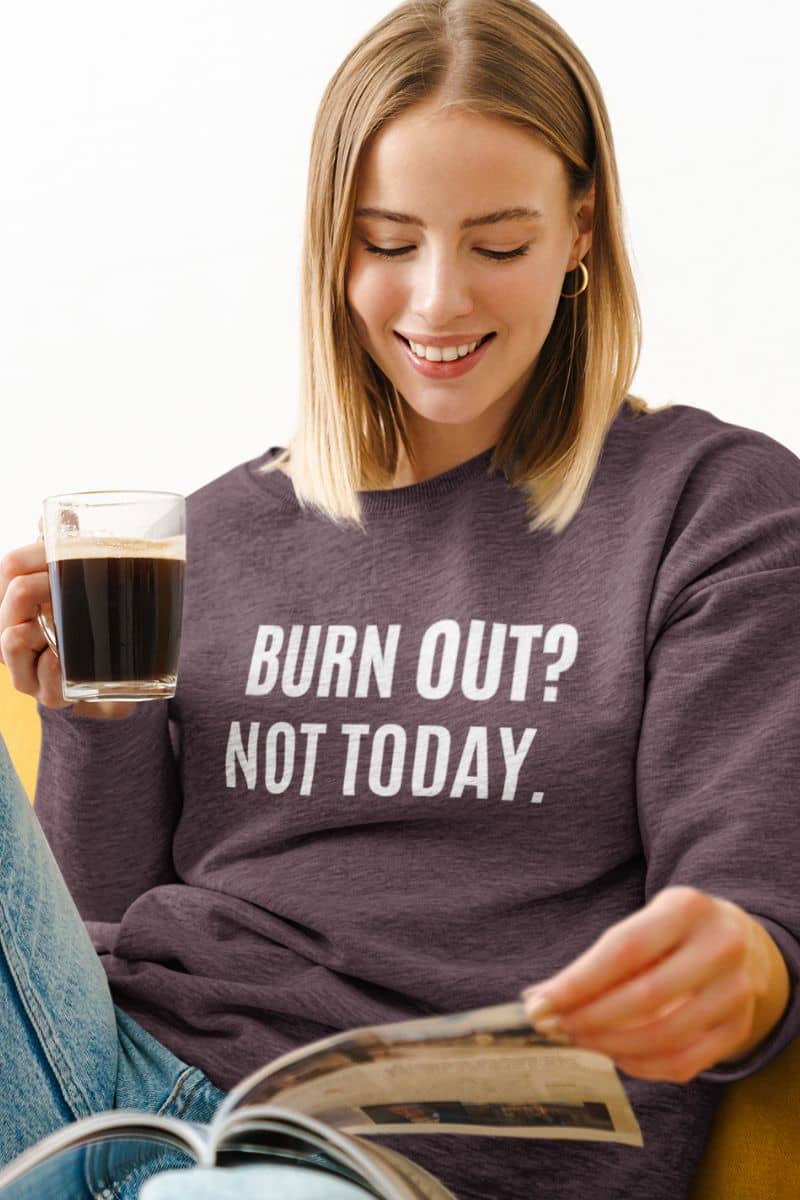 empowering crewneck for women with a woman wearing a sweatshirt with 'BURN OUT? NOT TODAY.' text, holding a coffee cup and reading a newspaper.