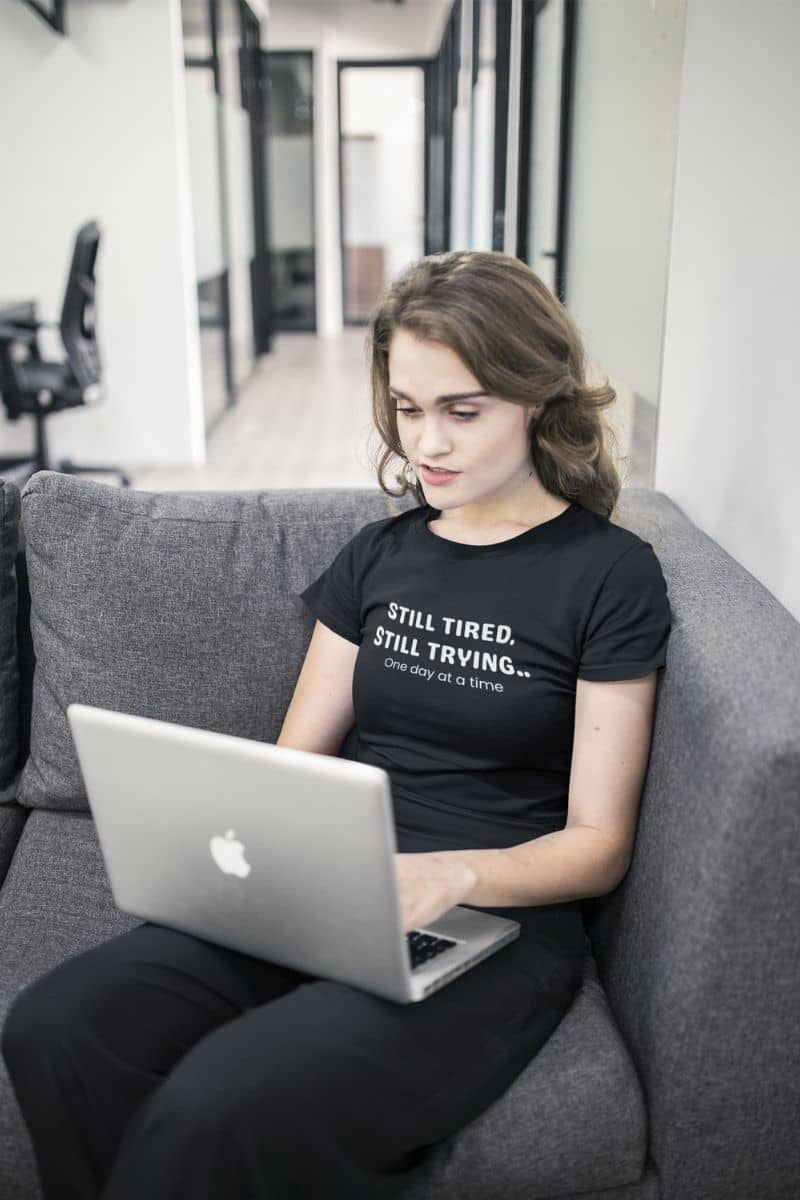 encouraging message t-shirt with a person sitting on a couch using a laptop with a black t-shirt that reads 'STILL TIRED. STILL TRYING.'