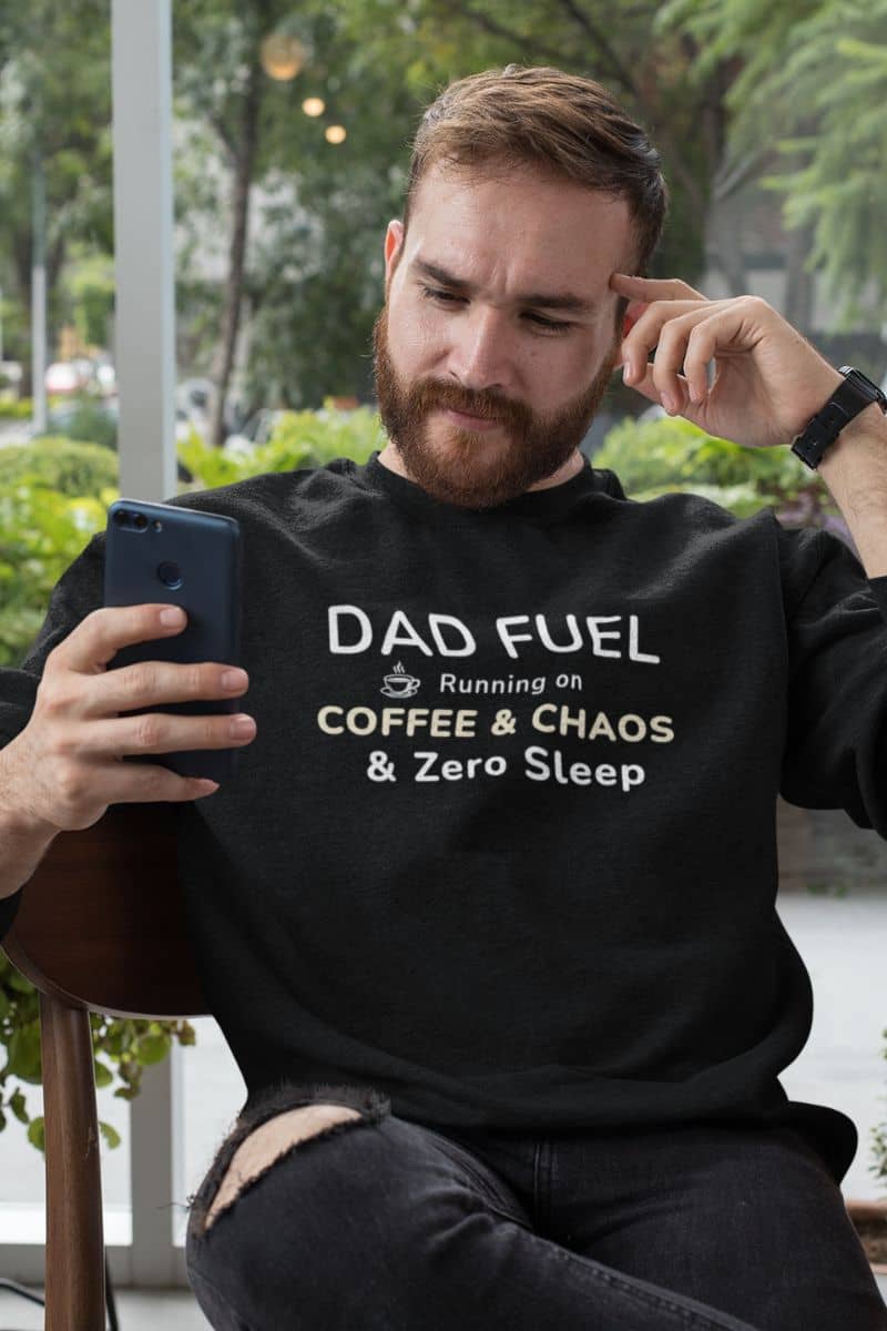 exhausted dad sweatshirt with a man wearing a black sweatshirt with text 'DAD FUEL – Running on Coffee & Chaos & Zero Sleep', sitting outdoors holding a phone.